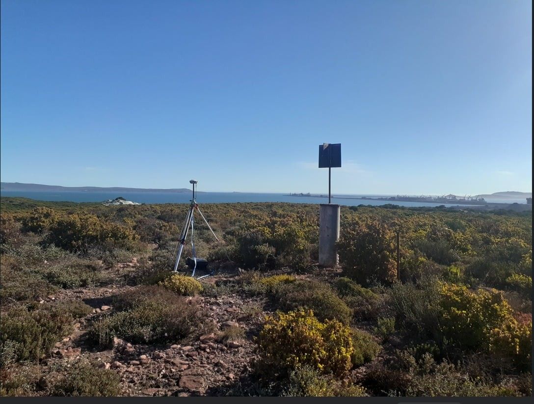 A GNSS receiver on a tripod in fynbos, with the sea in the distance