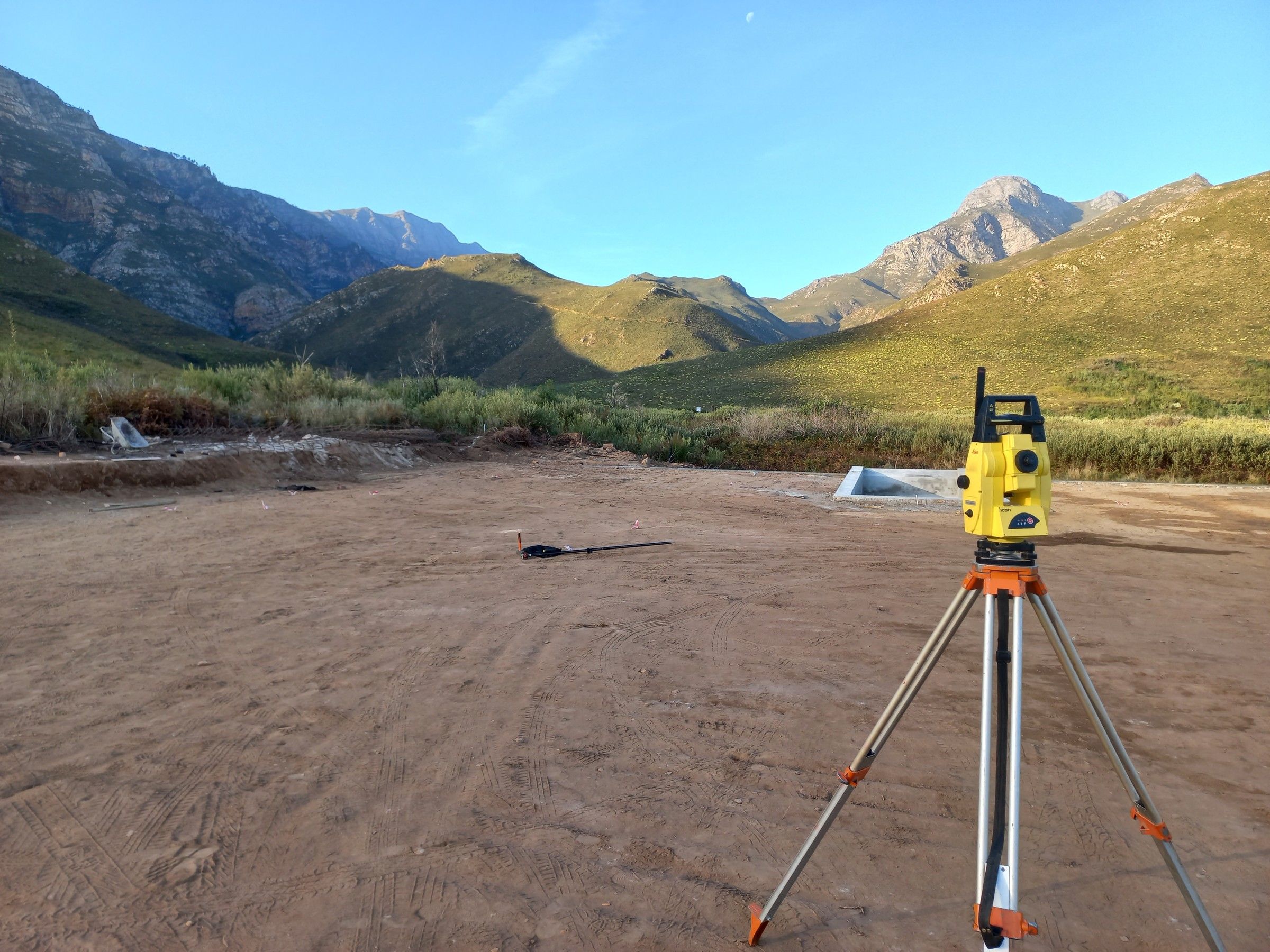 A total station on a tripod, set up on a levelled pad in a mountain valley